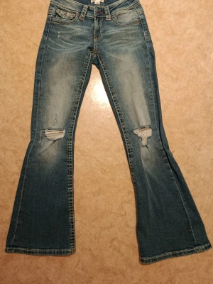 Blå bootcut jeans med slitningar - Snygga blå bootcut jeans med slitningar på knäna och lätt tvättad look. Jeansen har fem fickor och dekorativa sömmar på bakfickorna. Perfekta för en avslappnad och trendig stil. Är i storlek 158 cm men passar dom som har storlek XS-S