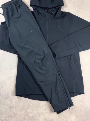 Nike unlimited  - Snyggt svart träningsset från Nike med matchande hoodie och byxor. Jackan har dragkedja, huva med dragsko och diskret Nike-logga på bröstet. Byxorna har resår i midjan, dragkedja vid benslut och liten Nike-logga. Perfekt för gymmet eller löprundan. Köpt för 2800 perfekt inför sommaren 