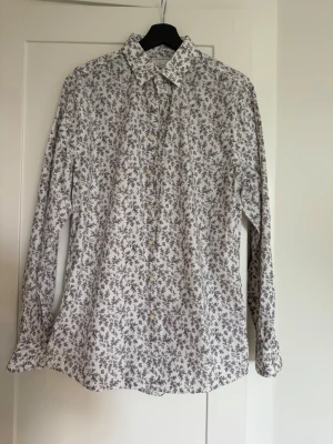 Vit blommig skjorta med knappar - Vit skjorta med små blommiga mönster i svart. Skjortan har klassisk krage, långa ärmar och knäppning framtill. Tillverkad i mjuk bomull som känns skön mot huden. Perfekt för dig som gillar en stilren men ändå unik look.
