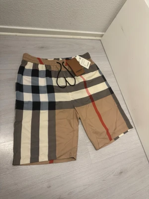 Burberry badbyxor i klassiskt rutmönster - Snygga badbyxor från Burberry i deras ikoniska rutmönster med färger i beige, svart, vitt, grått och rött. Resår i midjan med svart dragsko och innerfoder i mesh. Perfekta för stranden eller poolen, riktigt stilrena och trendiga. Riktigt sköna badbyxor. De är inte använda. Taggen sitter fortfarande kvar. Jag skickar samma dag eller dagen efter :)