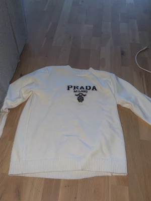 Vit stickad tröja från Prada - Vit stickad tröja från Prada med svart broderad logga och texten 'PRADA MILANO' på bröstet. Tröjan har rund halsringning, långa ärmar och ribbstickade muddar vid ärmslut och nederkant. Perfekt för dig som gillar exklusiva märken.