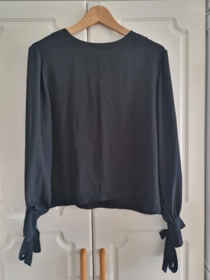 Svart blus med knytband vid ärmslut - Stilren svart blus med rund halsringning och långa ärmar som avslutas med snygga knytband vid handlederna. Blusen har en enkel knappstängning i nacken och är tillverkad i ett mjukt, lätt tyg som faller fint.