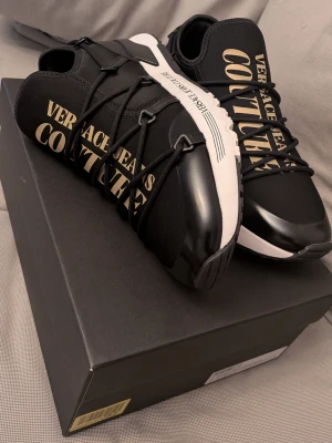 Svarta sneakers Versace Jeans Couture - PRIS KAN DISKUTERAS VID SNABB AFFÄR. .Stilrena svarta sneakers från Versace Jeans Couture med stora guldfärgade logotexter på sidan och ovandelen. Skorna har vita sulor, svarta snören och detaljer i både textil och syntetmaterial. Perfekta för dig som vill sticka ut med en lyxig streetwear-look.