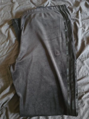 Svarta velourbyxor från Adidas - Oanvända, bara provat dom men gillade aldrig dom Mjukisbyxor i grå velour från Adidas med klassiska tre ränder längs benen och broderad logga på låret. Byxorna har en avslappnad passform och resår i midjan. Perfekta för chill dagar eller när du vill ha en sportig look.