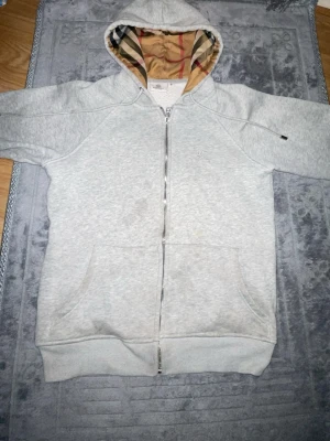 Grå zip hoodie från Burberry - Säljer en grå zip hoodie från Burberry med klassiskt rutigt foder i huvan. Hoodien har dragkedja framtill, två stora fickor och ribbade muddar. Perfekt för dig som gillar stilrena plagg med exklusiva detaljer.