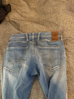 Blå Replay jeans med slitningar - Säljer ett par snygga blå jeans från Replay med ljusa slitningar och klassisk femficksdesign. Jeansen har en skinnpatch med logga bak i midjan och detaljerade sömmar på bakfickorna. Perfekta för en avslappnad och trendig look.