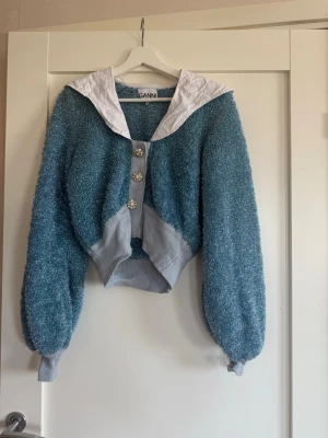 Blå glittrig kofta från Ganni - Blå, glittrig kofta från Ganni med stora dekorativa knappar och vit sjalkrage. Koftan har ballongärmar, ribbade muddar och en croppad passform. En liten sminkfläck på kragen (se bild) + en knapp som hänger löst (se bild). Står XS men passar S.
