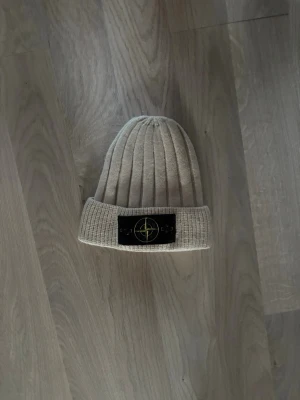 Beige mössa från Stone Island - Säljer en beige mössa från Stone Island med uppvikt kant och den klassiska svarta logotypen framtill. Mössan är tillverkad i ett mjukt och stretchigt material som sitter skönt på huvudet. Perfekt för dig som vill ha en stilren och trendig accessoar. Ny utan prislapp!