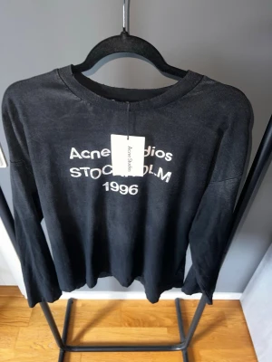 Svart långärmad topp Acne Studios - Svart långärmad topp från Acne Studios med vit text 'Acne Studios STOCKHOLM 1996' framtill. Klassisk rund halsringning och avslappnad passform. Perfekt för en stilren och trendig look.