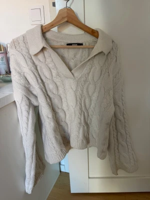 Beige kabelstickad tröja från Bik Bok - Kabelstickad tröja i beige från Bik Bok med v-ringning och bred krage. Tröjan har långa ärmar och avslappnad passform, perfekt för lager-på-lager. Mjukt material och klassiskt flätmönster ger en trendig och mysig look.