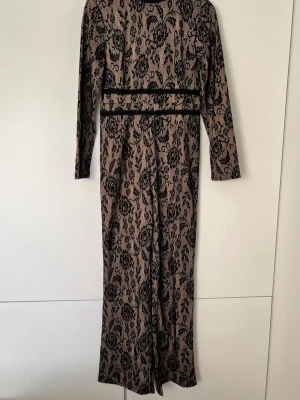 Beige och svart mönstrad jumpsuit - Snygg jumpsuit i beige med svart blommigt spetsmönster. Långärmad modell med markerad midja och vida ben. Dragkedja i ryggen för enkel påklädning. Perfekt för dig som vill sticka ut med en unik och trendig look.