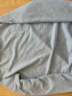 Grå sweatshirt från Moncler - Snygg grå sweatshirt från Moncler med broderad logga på bröstet och patch på ärmen. Tröjan har rund halsringning och är tillverkad i mjukt bomullsmaterial. Perfekt för en avslappnad och stilren look.