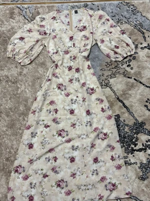 Blommig långklänning med puffärm - Romantisk långklänning med blommigt mönster i lila och rosa toner på en ljus botten. Klänningen har v-ringning, markerad midja och puffiga trekvartsärmar. Perfekt för sommarens alla tillfällen.