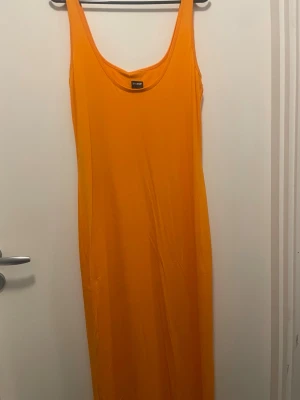 Orange långklänning från Gina Tricot - Snygg orange långklänning från Gina Tricot i modellen Maya dress. Klänningen är ärmlös med rund halsringning och har en tight passform. Perfekt för sommarens alla tillfällen. Materialet är mjuk och stretchig polyester.