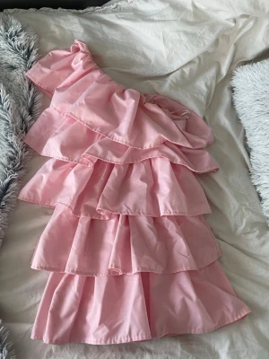 Rosa volangklänning från Anielle - Supersöt rosa klänning från Anielle med flera lager volanger och oneshoulder-design. Klänningen har en kort längd och är perfekt för dig som vill sticka ut med en feminin och trendig look. Tillverkad i ett mjukt tyg som känns skönt mot huden. Perfekt som suck klänning eller midsommarklänning