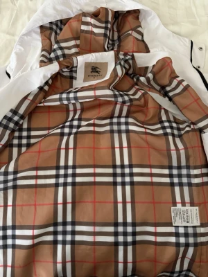Vit vindjacka från Burberry - Snygg vit vindjacka från Burberry med klassiskt rutigt foder i beige, svart, vitt och rött. Jackan har huva, svarta dragkedjor och detaljer samt Burberry-logga på bröstet. Perfekt för en trendig och stilren look.