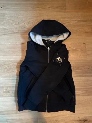 Svart hoodie från Moose Knuckles - Svart hoodie med dragkedja från Moose Knuckles. Utrustad med huva och mjukt vitt foder på insidan. Har en ikonisk logga i metall på ärmen och två fickor framtill. Perfekt för en chill och stilren look. Pris går att diskutera lite!