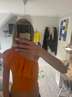 Orange one shoulder topp med volang - Trendig orange topp med one shoulder-design och stor volang längs axeln. Toppen är croppad och har en tight passform med ribbad struktur i tyget. Perfekt för dig som vill sticka ut med en färgstark och unik stil.