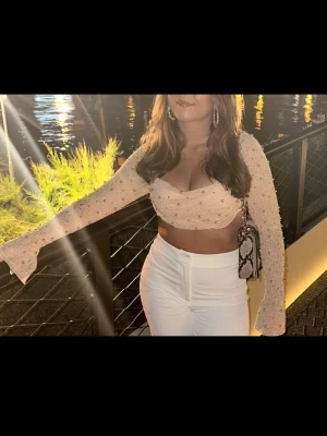 Beige croppad topp med pärlor från Oh Polly - Superfin croppad topp i beige med långa ärmar och pärldetaljer över hela tyget. Toppen har en söt v-ringning och är figurnära med lätt transparent material. Perfekt för dig som vill sticka ut lite extra.