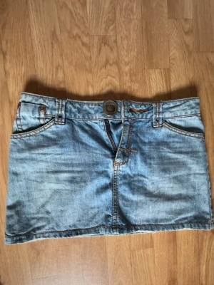 Ljusblå jeanskjol med fickor - Snygg ljusblå jeanskjol i klassisk denim med fem fickor, bälteshällor och knappstängning framtill. Kjolen har en kort modell och coola slitningar för en avslappnad look. Perfekt att matcha med sneakers eller boots.
