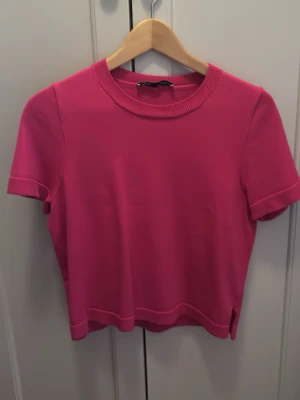 Rosa topp från Zara - Snygg rosa topp från Zara med rund halsringning och korta ärmar. Toppen har ribbade detaljer vid halsen och är tillverkad i ett mjukt material som känns skönt mot huden. Perfekt att styla med jeans eller kjol för en trendig look.