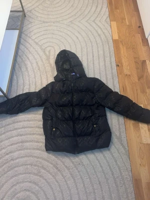 Svart pufferjacka med huva - Säljer en svart gucci pufferjacka med huva och dragkedja framtill. Jackan har två fickor med knappar och ett diskret mönster över hela tyget. Perfekt för kalla dagar och riktigt skön att bära. Passar dig som gillar en stilren och trendig look.