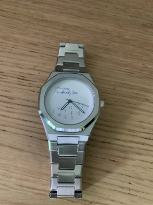  klocka  - Säljer en unik klocka i silverfärgat metallarmband med vit urtavla. På urtavlan står det 'Who cares I'm already late' och siffrorna är utspridda på ett lekfullt sätt. Perfekt accessoar för dig som gillar humor och stilren design.