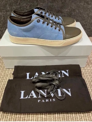 Lanvin blå - Snygga sneakers från Lanvin med blå mocka på sidorna. Klassisk låg modell med svarta skosnören och diskreta metalldetaljer. Perfekta nu till sommaren med lite ljusare färger!