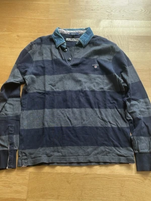 Randig långärmad skjorta från Gant - Snygg långärmad skjorta från Gant med breda marinblå och grå ränder. Skjortan har en klassisk krage i denim, knappar vid halsen och ribbade muddar vid ärmsluten. Perfekt för en avslappnad men stilren look.