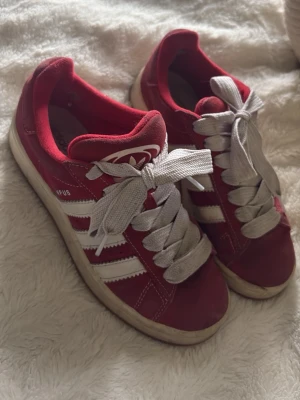 Röda Adidas Campus sneakers - Snygga röda Adidas Campus sneakers med klassiska vita ränder på sidorna och breda vita skosnören. Ovandelen är i mocka och sulan är vit med gummidetaljer. Perfekta för en sportig och trendig stil.