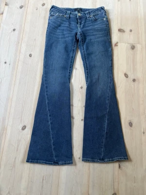 Joey True Religion jeans - Snygga blå bootcut jeans från True Religion med klassiska kontrastsömmar och dekorativa bakfickor med lock och knapp. Jeansen har låg midja och utsvängda ben, perfekt för en trendig och avslappnad look.