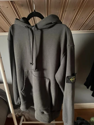 Mörkgrå hoodie från Stone Island - Snygg mörkgrå hoodie från Stone Island med klassisk patch på vänster ärm. Hoodien har huva med dragsko och en stor magficka framtill. Perfekt för dig som gillar streetwear och vill ha en stilren look.