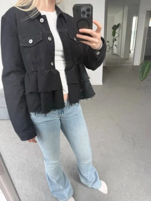 Svart volang jeansjacka - Säljer en svart jeansjacka med volangdetaljer och råa kanter nedtill. Jackan har två bröstfickor med knappar och klassisk krage. Perfekt för dig som vill ha en unik twist på den klassiska jeansjackan.