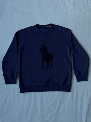 Mörkblå stickad tröja från Polo Ralph Lauren - Mörkblå stickad tröja från Polo Ralph Lauren med stort ton-i-ton broderat polospelar-motiv på bröstet. Tröjan har rund halsringning, långa ärmar och ribbade muddar vid ärmslut och nederkant. Perfekt för en stilren och klassisk look.