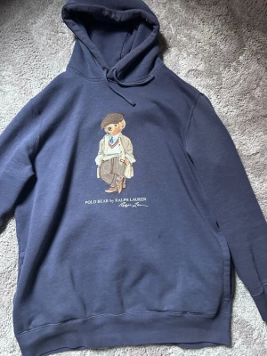 Blå Polo Bear hoodie från Ralph Lauren - Mörkblå hoodie från Ralph Lauren med den ikoniska Polo Bear-trycket på bröstet. Hoodien har huva med dragsko och ribbade muddar. Tillverkad i mjukt bomullsmaterial.( Köptes 2799kr)