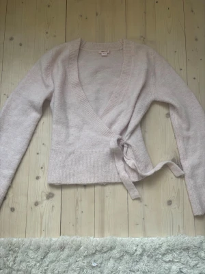 Ljusrosa omlottkofta med knytband - Superfin ljusrosa omlottkofta med knytband i midjan och djup v-ringning. Koftan har långa ärmar och bred ribbad kant nedtill. Perfekt att slänga på sig över ett linne eller t-shirt för en chill look.