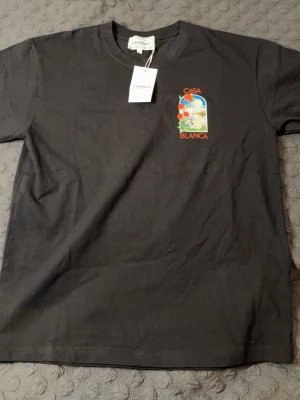 Svart Casa Blanca t-shirt med tryck - Svart t-shirt från Casa Blanca med färgglatt tryck av rosor och landskap både fram och bak. Klassisk passform med rund hals och korta ärmar. Perfekt för dig som vill sticka ut med en snygg och unik design. STORLEK M, PASSAR L-XL. HELT NY, OANVÄND