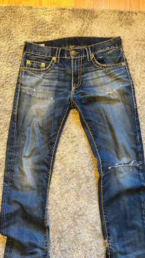 Vintage true religion jeans - Säljer ett par blå jeans med tydliga slitningar och kontrastsömmar. Jeansen har fem fickor, dekorativa nitar och en amerikansk flagga på linningen. Snyggt tvättade detaljer och en avslappnad passform gör dem perfekta till en cool streetstil.