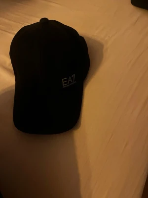 Svart keps från EA7 Emporio Armani - Snygg svart keps från EA7 Emporio Armani med broderad logga framtill och justerbar rem bak. Klassisk böjd skärm och stilren design, perfekt för en trendig streetwear-look. Tillverkad i mjukt och bekvämt material.
