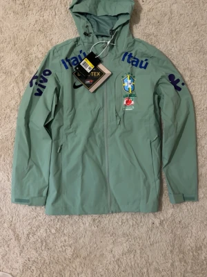 Nike Brasil vindjacka GORE-TEX grön  - Snygg grön vindjacka från Nike med Brasilien CBF-logga och sponsormärken som Itaú och Vivo. Jackan är tillverkad i GORE-TEX-material, har huva och dragkedja framtill samt fickor med dragkedja. Perfekt för dig som gillar sportig stil och vill ha något unikt.