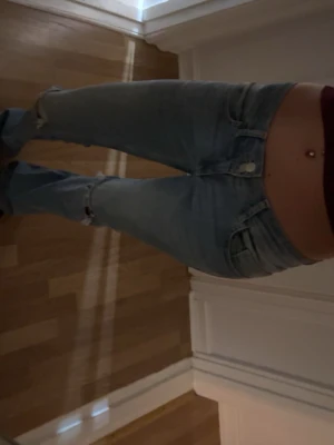 Ljusblå slitna bootcut jeans - Säljer ett par ljusblå bootcut jeans med slitningar vid knäna och klassiska fem fickor. Jeansen har låg midja och stängs med knapp och dragkedja. Perfekta för en avslappnad och trendig look.
