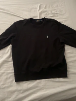 Svart sweatshirt från Polo Ralph Lauren - Klassisk svart sweatshirt från Polo Ralph Lauren med rund halsringning och den ikoniska broderade loggan på bröstet. Tröjan har en enkel och stilren design, perfekt för en avslappnad look. Tillverkad i mjukt material för extra komfort.