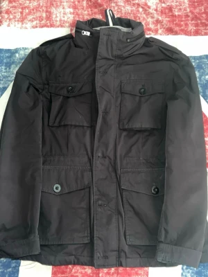 Svart overshirt från Berkeley. - Stilren svart overshirt med fyra stora fickor framtill och hög krage. Jackan har en rak passform och stängs med dragkedja och knappar. Perfekt för blåsiga dagar och enkel att matcha med olika outfits.