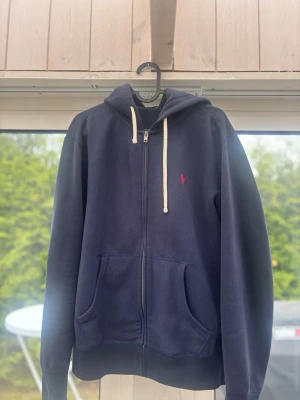 Polo Ralph Lauren hoodie – marinblå klassiker 💙 - Säljer en snygg zip-hoodie från Polo Ralph Lauren i en tidlös marinblå färg. Perfekt basplagg som funkar till allt – både till vardags och mer uppklädda streetwear-looks.  Tröjan har den ikoniska röda hästen broderad på bröstet och vita dragsnören som ger en snygg kontrast. Full zip-modell med huva och två framfickor, vilket gör den både stilren och praktisk.  Mjuk och bekväm kvalitet som håller formen bra. Passar perfekt för lager-på-lager eller som ensam hoodie