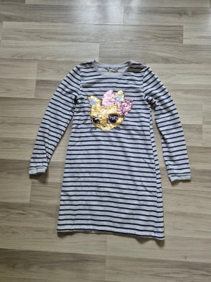 Randig klänning med paljettmotiv H&M - Grå- och vitrandig långärmad klänning från H&M med ett stort paljettmotiv av ett djuransikte med rosett framtill. Mjuk och bekväm bomullskvalitet, perfekt för en lekfull stil.