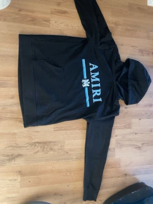 Amiri hoodie - Svart hoodie från Amiri med stort blått och vitt logotryck på bröstet. Klassisk känguruficka framtill och huva. Mjuk bomullskänsla och avslappnad passform, perfekt för en cool streetwear-look. Står M men passar som L