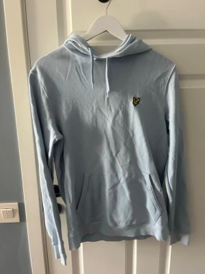 Ljusblå hoodie från Lyle & Scott - Snygg ljusblå hoodie från Lyle & Scott med klassisk gul logga på bröstet. Hoodien har huva med dragsko, magficka och ribbade muddar vid ärmslut och nederkant. Perfekt för en chill och avslappnad stil.