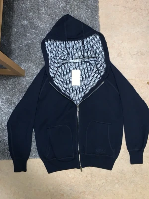 L. Dior kofta/cardigan  - Säljer denna kofta/cardigan för att har bytt ny stil till casual/Classic har inte plats för nya stilen i garderoben därför väljer och sälja Snygg marinblå hoodie från Christian Dior med dragkedja och två stora fickor framtill. Insidan av huvan har ett ikoniskt Dior-mönster i svart och vitt. Tillverkad i Italien och gjord i mjukt material som känns lyxigt och bekvämt. Perfekt för dig som vill ha något exklusivt och stilrent.