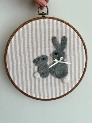 Broderade tavlor med ullkaniner - Unik broderade tavla med två små gråa ullkaniner, monterad i en rund ram. Motivet är gjort på beige- och vitrandigt tyg och dekorerat med vit rosett. Perfekt som gullig väggdekoration till barnrum eller present.