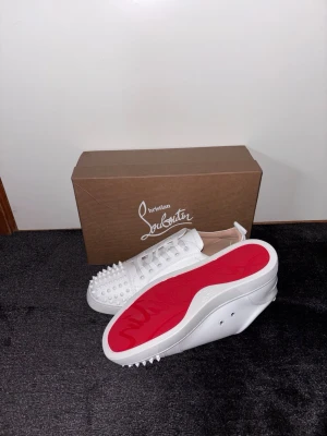Vita Christian Louboutin sneakers med nitar - Snygga vita sneakers från Christian Louboutin med ikonisk röd sula och coola nitar framtill. Skorna har lågt skaft och är tillverkade i läder för en lyxig känsla. Perfekta för dig som vill sticka ut med en exklusiv och edgy stil.
